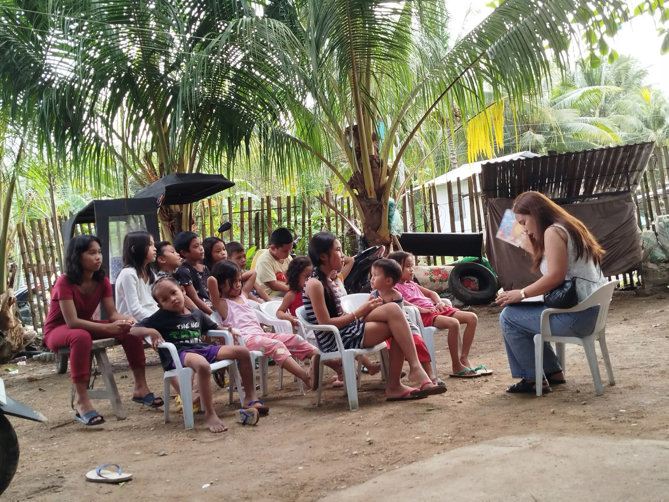 Children seated outdoors while a teacher speaks with them